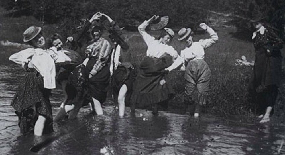 women with their skirts tucked up wading in a stream