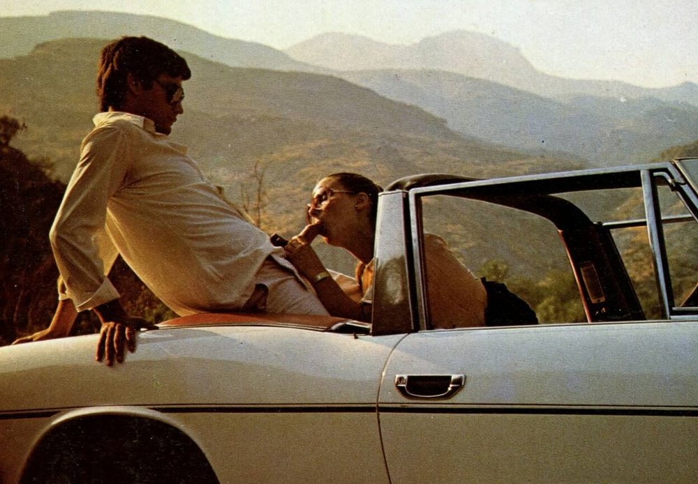 A man sits on the top of the seats in the back of his convertible and braces himself against the trunk lid while his cute woman gives him a loving blowjob as the summer sun sets over the mountains