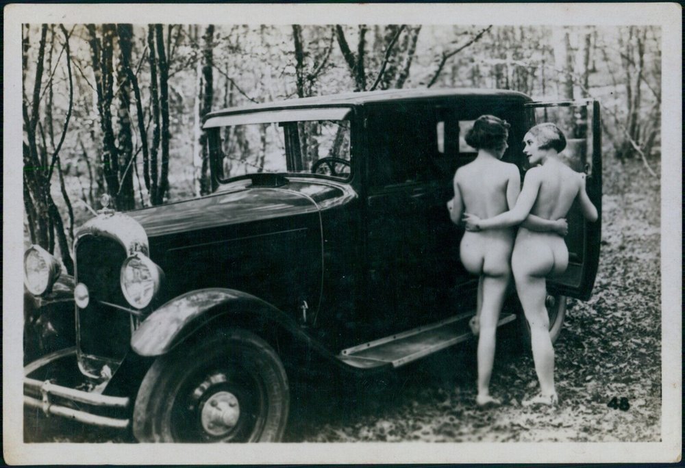 two nude women climbing into the back seat of an old-fashioned touring car parked in an isolated forest